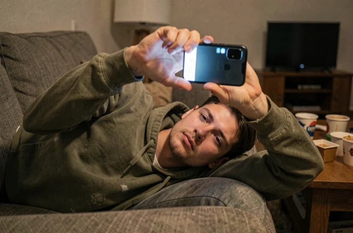 Jeune homme allongé sur un canapé, absorbé par son smartphone dans une atmosphère de surconsommation numérique à domicile